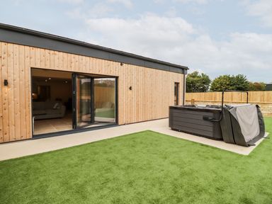 A patio with artificial grass a hot tub with a partially open cover and a wooden exterior house with glass doors at Bears Court Barn 3 Little Rissington near Bourton-On-The-Water