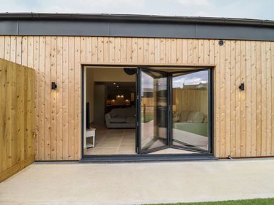 A wooden exterior with folding glass doors leading to a living area with sofas and kitchen at Bears Court Barn 3 in Little Rissington near Bourton-On-The-Water
