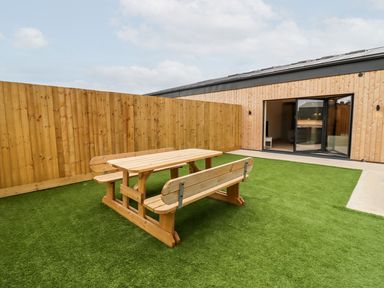 An outdoor area with a wooden picnic table on artificial grass next to a wooden building with sliding glass doors at Bears Court Barn 3 Little Rissington near Bourton-On-The-Water