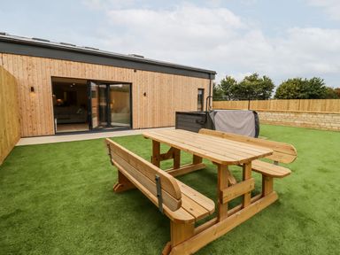 An outdoor area with artificial grass a wooden picnic table and benches a hot tub and a wooden building with open glass doors at Bears Court Barn 3 Little Rissington near Bourton-On-The-Water