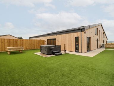 A backyard with artificial grass wooden picnic table hot tub and a wooden building at Bears Court Barn 3 Little Rissington near Bourton-On-The-Water