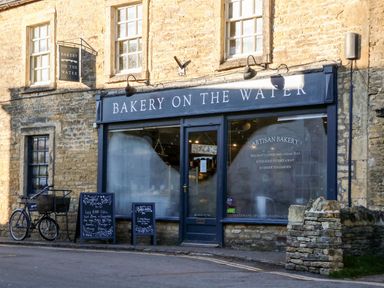 The exterior of a bakery with a sign bakery on the water and a bicycle outside at Bears Court Barn 3 Little Rissington near Bourton-On-The-Water