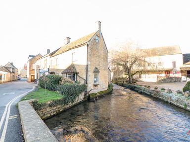 A stone building next to a flowing river with a small road and other buildings nearby at Bears Court Barn 3 Little Rissington near Bourton-On-The-Water