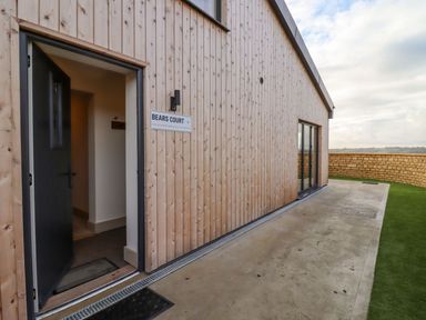 Exterior view of a wooden building with an open black door and a sign that reads Bears Court 4 at Little Rissington near Bourton-On-The-Water