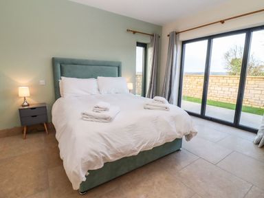 A bedroom with a double bed with white bedding and towels on it two bedside tables with lamps and sliding glass doors at Bears Court Barn 4 in Little Rissington near Bourton-On-The-Water