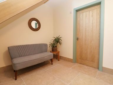 A seating area with a gray upholstered bench a wooden side table with a potted plant a round wall mirror and a wooden door with light blue trim at Bears Court Barn 4 Little Rissington near Bourton-On-The-Water