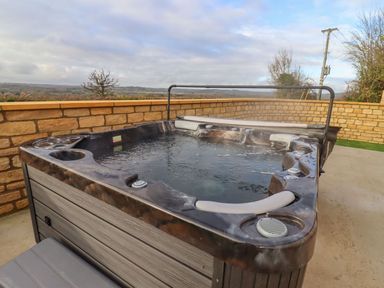 An outdoor hot tub on a concrete patio surrounded by a stone wall with a view over countryside at Bears Court Barn 4 Little Rissington near Bourton-On-The-Water
