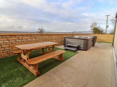 An outdoor patio with a wooden picnic table and benches and a hot tub with steps behind a low stone wall at Bears Court Barn 4 Little Rissington near Bourton-On-The-Water