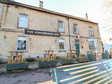 The exterior of The Kingsbridge Inn with wooden tables and planters outside and a green bench in front