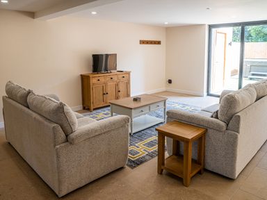 A living room with two sofas a coffee table a wooden cabinet with a television and sliding glass door at Bears Court Barn 7 Little Rissington near Bourton-On-The-Water
