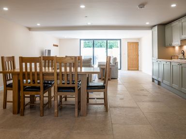 An open living space with a wooden dining table and chairs a seating area with sofas large windows and kitchen cabinets at Bears Court Barn 7 Little Rissington near Bourton-On-The-Water