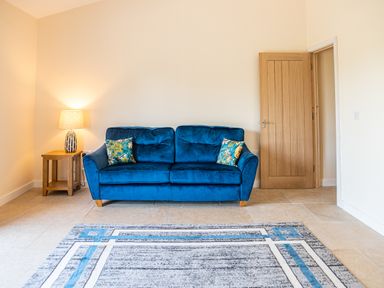 A blue sofa with patterned cushions next to a wooden side table with a lamp in a room with a wooden door and a patterned rug on the floor at Bears Court Barn 7 Little Rissington near Bourton-On-The-Water
