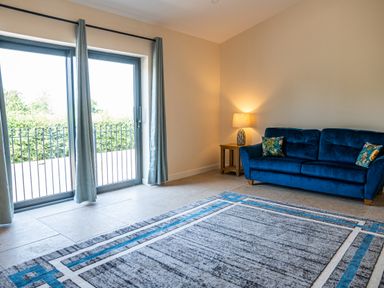 A living room with a blue sofa two cushions a side table with a lamp carpet and sliding glass door at Bears Court Barn 7 in Little Rissington near Bourton-On-The-Water