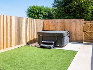 A backyard with a hot tub on a concrete slab bordered by artificial grass and wooden fencing at Bears Court Barn 7 Little Rissington near Bourton-On-The-Water