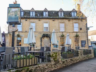 An outdoor seating area with tables and closed umbrellas in front of a stone building with multiple windows and a sign reading Old Manse Hotel