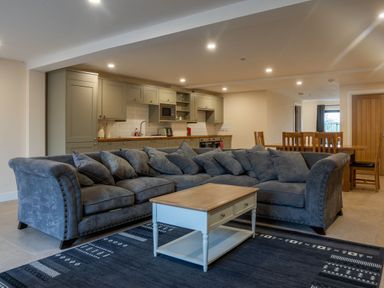 An open plan room with a grey sectional sofa coffee table kitchen cabinets and a dining table with chairs at Bears Court Barn 8 Little Rissington near Bourton-On-The-Water