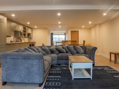 An open plan living room with a grey sectional sofa a white coffee table a dark patterned rug a dining table with wooden chairs and a kitchen with grey cabinets at Bears Court Barn 8 in Little Rissington near Bourton-On-The-Water