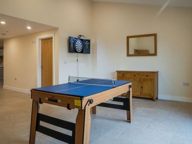 A game room with a ping pong table a dartboard mounted on the wall a wooden door a wooden cabinet with drawers and a mirror above it at Bears Court Barn 8 Little Rissington near Bourton-On-The-Water