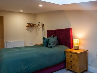 A bedroom with a bed with a purple headboard teal bedding a wooden nightstand with a lamp and a wall mounted clothes rail with hangers at Bears Court Barn 8 Little Rissington near Bourton-On-The-Water