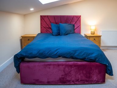 A bedroom with a bed with a red headboard and blue bedding with wooden bedside tables and a lamp at Bears Court Barn 8 in Little Rissington near Bourton-On-The-Water