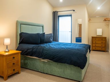 A bedroom with a bed with dark bedding a bedside table with lamp a mirror and wooden chest of drawers at Bears Court Barn 8 in Little Rissington near Bourton-On-The-Water