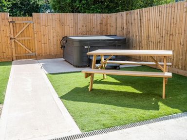 A backyard with synthetic grass a wooden picnic table a hot tub and a wooden fence at Bears Court Barn 8 Little Rissington near Bourton-On-The-Water