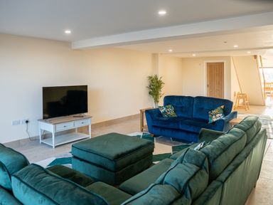 A living room with green and blue sofas a television on a white stand and a plant at Bears Court Barn 5 in Little Rissington near Bourton-On-The-Water