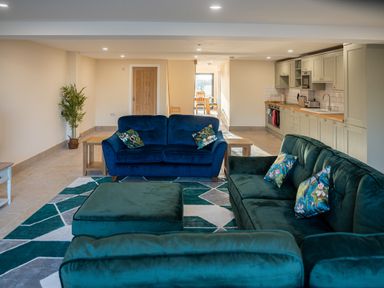 An open-plan living room with blue and green sofas a patterned rug wooden side tables a kitchen area with cabinets and appliances and a dining area in the background at Bears Court Barn 5 in Little Rissington near Bourton-On-The-Water