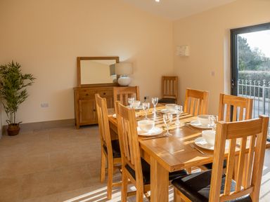 A dining room with a wooden table set for six with bowls and glasses a wooden sideboard with a lamp a plant in the corner and a glass door letting in natural light at Bears Court Barn 5 Little Rissington near Bourton-On-The-Water