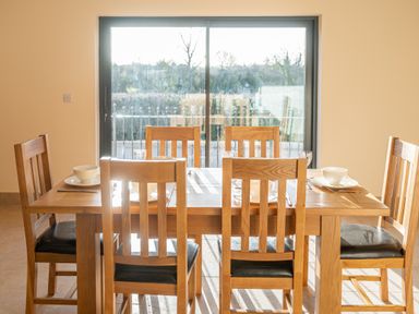 A dining room with a wooden table and six chairs set with bowls, plates, and glasses in front of a large window at Bears Court Barn 5 in Little Rissington near Bourton-On-The-Water