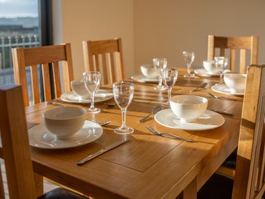 A wooden dining table set with white bowls, plates, forks, knives, spoons, and clear wine glasses at Bears Court Barn 5 Little Rissington near Bourton-On-The-Water