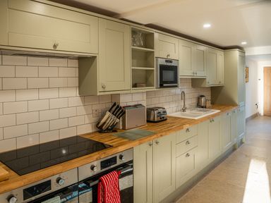 A kitchen with wooden countertops, green cabinets, a stovetop, oven, knife set, toaster, microwave, sink, and tiled backsplash at Bears Court Barn 5 in Little Rissington near Bourton-On-The-Water