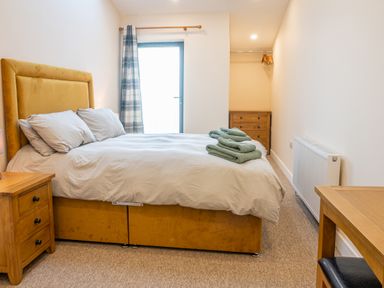 A bedroom with a double bed with yellow headboard white bedding bedside tables a dresser towels on bed and a window at Bears Court Barn 5 in Little Rissington near Bourton-On-The-Water