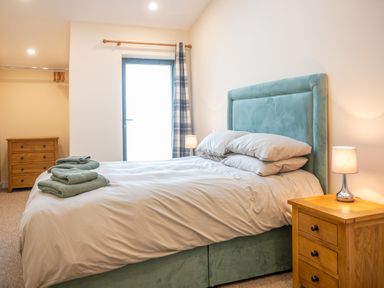 A bedroom with a bed with pillows and towels a bedside table with a lamp a chest of drawers and a window with a curtain at Bears Court Barn 5 in Little Rissington near Bourton-On-The-Water