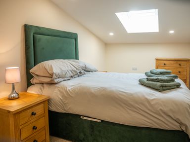 A bedroom with a bed with green headboard and folded towels on it a wooden nightstand with a lamp and a wooden dresser at Bears Court Barn 5 Little Rissington near Bourton-On-The-Water