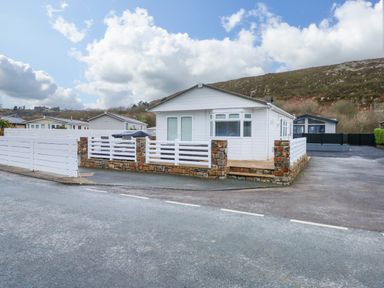 A white single-story house with a stone and white fence on a corner lot with a paved driveway and hills in the background at Un Diwrnod (One Day) The Warren T22 The Warren near Abersoch