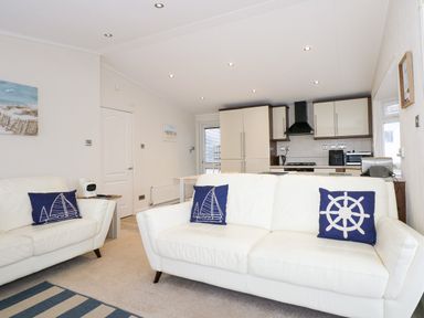 A living room with white sofas and blue nautical pillows facing a kitchen with cream cabinets at Un Diwrnod The Warren T22 The Warren near Abersoch