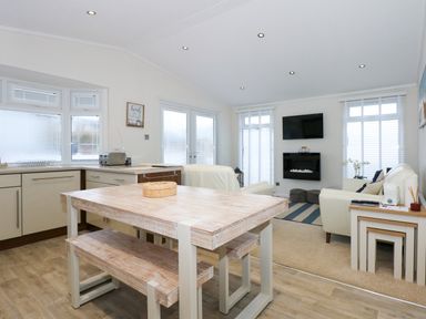 A kitchen area with wooden dining table and bench seats next to a living area with white leather sofas and a wall-mounted TV at Un Diwrnod The Warren T22 The Warren near Abersoch