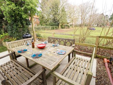 An outdoor wooden dining table set with plates glasses and a bowl on a patio next to a grassy fenced garden at Natterjacks in Syderstone