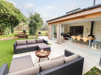 An outdoor patio with two dark wicker sofas and a fire pit next to a garden and an open doorway leading to a dining area with a table and chairs at Natterjacks in Syderstone