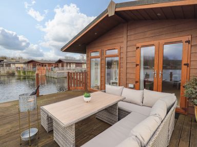 An outdoor deck with a wicker sectional sofa and table next to a wooden cabin by the water at Otters Reach near Carnforth