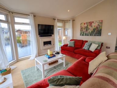 A living room with red sofas a white coffee table a wall-mounted tv and large windows overlooking water at Otters Reach near Carnforth