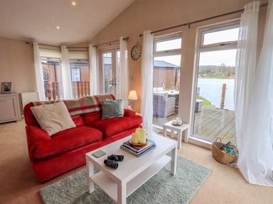A living room with a red sofa white coffee table and large windows overlooking a deck and lake at Otters Reach near Carnforth