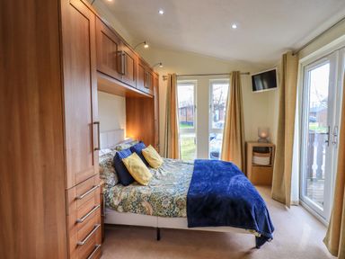 A bedroom with a bed, built-in wooden wardrobes, curtains, a small cabinet, wall-mounted tv, and glass doors at Otters Reach near Carnforth