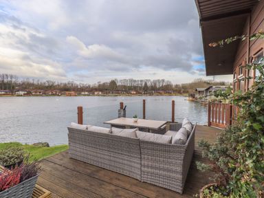 A patio with wicker seating and a table overlooking a lake with houses on the opposite shore at Otters Reach near Carnforth