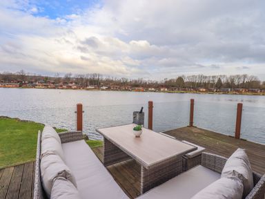 An outdoor seating area with a table and sofas on a wooden deck overlooking a lake with houses across the water at Otters Reach near Carnforth