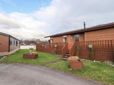 A brown wooden cabin with a fenced porch and planters on grass at Otters Reach near Carnforth
