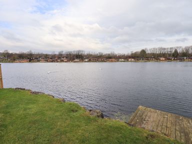 A lakeside view with wooden cabins across the water and a grassy shore with a wooden deck at Otters Reach near Carnforth
