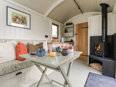 A small living space with a foldable table set with cups and flowers a built in bench with cushions a wood burning stove and a kitchen area at Blackthorn in South Leigh near Witney