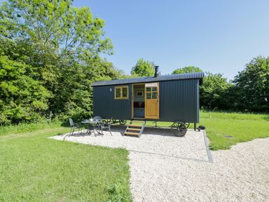 A small black corrugated structure with a wooden door and window on a gravel area with metal table and chairs surrounded by grass and trees at Blackthorn in South Leigh near Witney